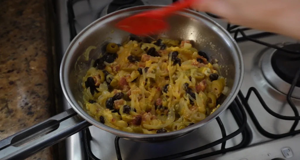 bacon, manteiga, cebola, ovos, azeitona e uva passa cozinhando em uma frigideira de inox